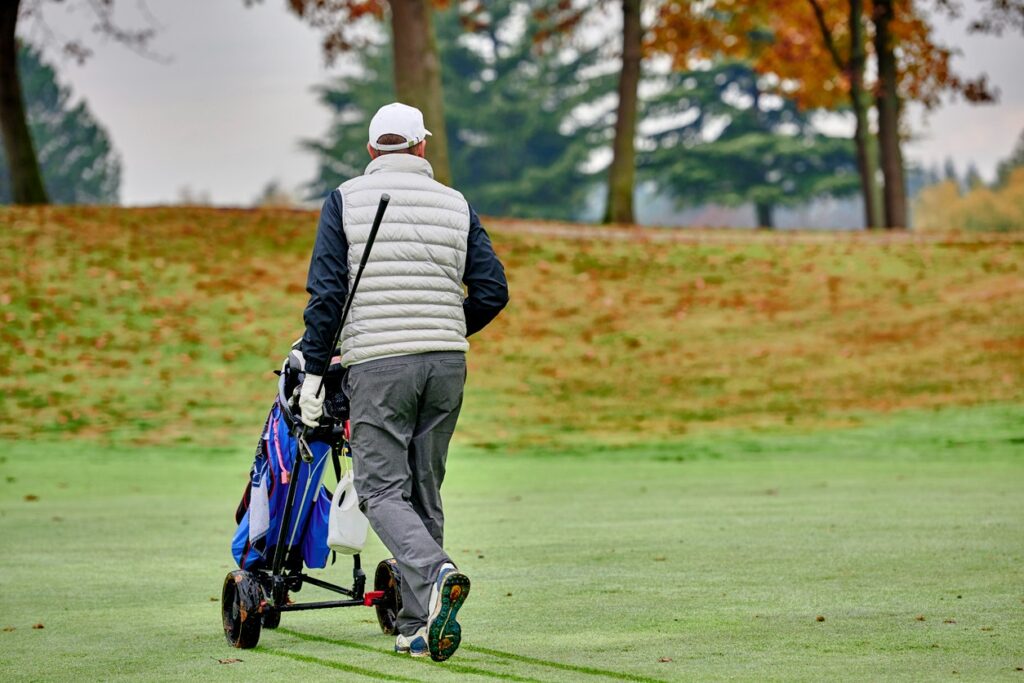 A man walks on a golf course with his golf trolley in fall weather.