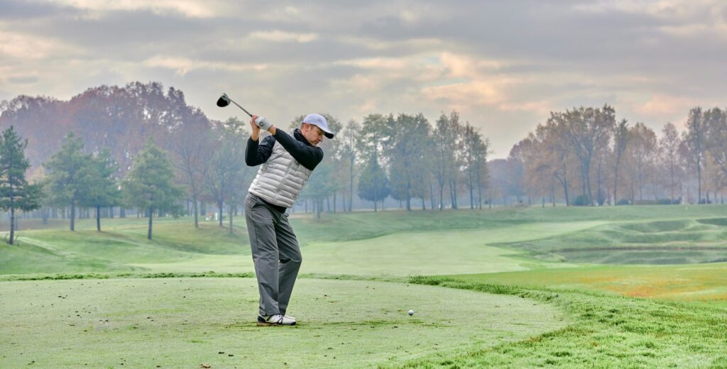 A male golfer swings while playing golf in cold weather.