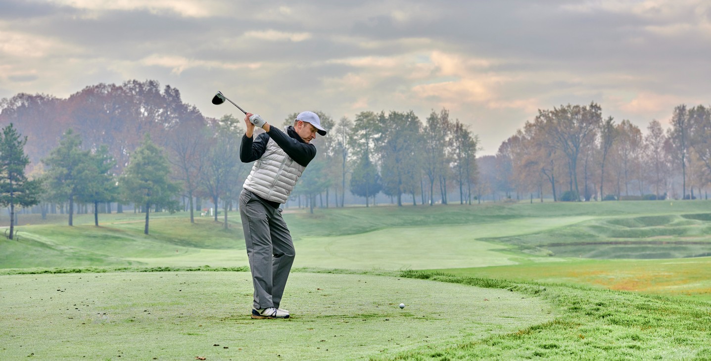 A male golfer swings while playing golf in cold weather. 